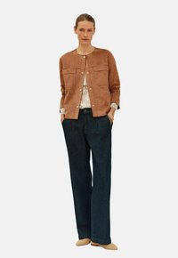 Giacca in suede tan con due tasche frontali, scollatura rotonda e chiusure a scatto, abbinata a jeans scuri a gamba larga e scarpe beige.