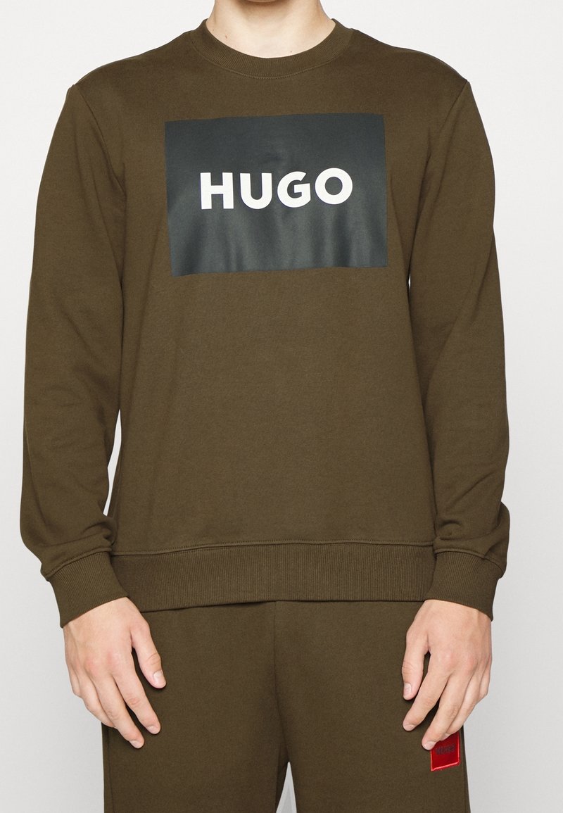 Personne portant un sweat-shirt vert olive et un pantalon assorti, le sweat-shirt comporte un rectangle noir avec le texte blanc "HUGO" sur la poitrine.