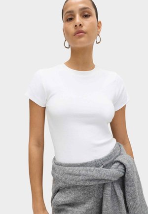 Ung kvinde med kort hår iført en hvid tætsiddende t-shirt og en grå sweater bundet om livet mod en ensfarvet baggrund.