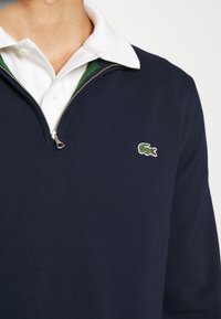 Marineblå strikket zip-sweater med hvid krave og grønne detaljer, der har et lille broderet logo på venstre bryst.