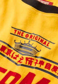 Camiseta de algodón amarilla con cuello acanalado negro y gráficos impresos en negrita, incluyendo "EL ORIGINAL" y un diseño de corona.