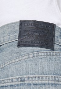 Jeans de mezclilla azul claro con un parche de cuero negro que presenta texto e imágenes en relieve, mostrando costuras detalladas y una textura suave.