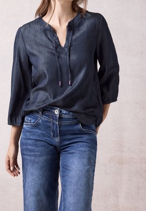 Femme portant un chemisier bleu foncé à manches longues avec des liens devant et un jean bleu en denim, debout devant un fond texturé clair.