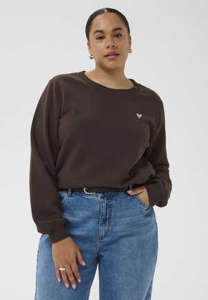 Žena v tmavě hnědé mikině s malým růžovým srdcem, zastrčené do modrých džínů s vysokým pasem a černým páskem, ruce volně položené.