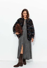 Veste en tricot avec des motifs bleus et marron, robe en denim gris à boutons, sac en suède marron avec franges et bottes de cowboy marron foncé.