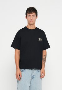 T-shirt nera in cotone con maniche corte, scollatura rotonda e un piccolo logo stampato sul lato sinistro del petto. Indossata con pantaloni corti in denim azzurro.