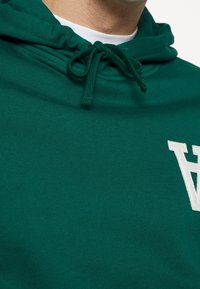 Zielony pulower z kapturem wykonany z miękkiego materiału, z kapturem na sznurku i białym logo na piersi o gładkim, teksturowanym wykończeniu.