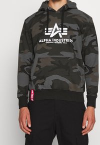 Kamouflage hoodie i mörkgrön och svart med vitt logotyp "Alpha Industries." Har en framficka och en kontrasterande röd etikett vid midjan.