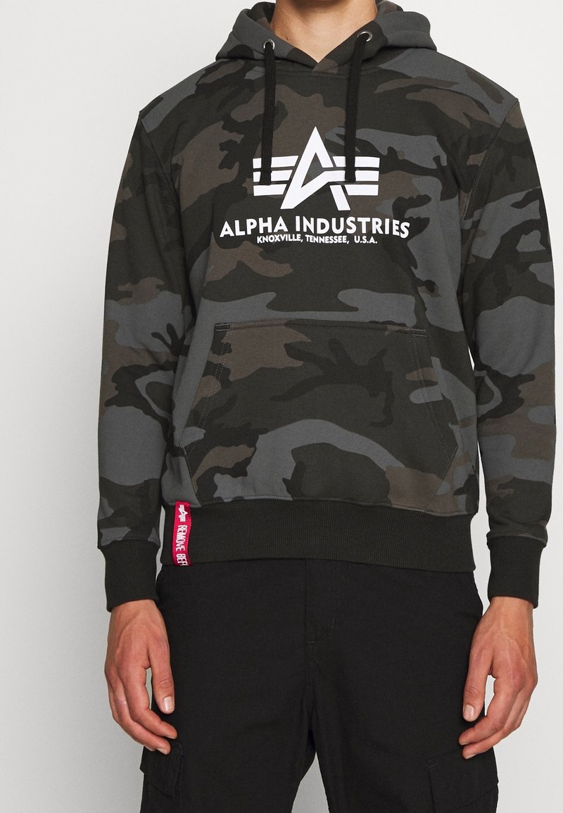 Kamouflage hoodie i mörkgrön och svart med vitt logotyp "Alpha Industries." Har en framficka och en kontrasterande röd etikett vid midjan.