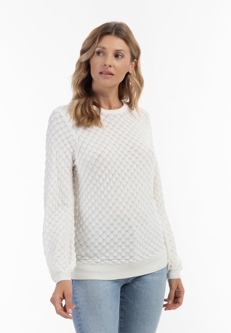 Maglione bianco strutturato con un motivo a rombi, maniche lunghe, polsini e orlo a coste, indossato con jeans azzurri chiari, su uno sfondo bianco.