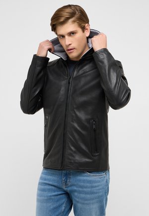 Junger Mann richtet die Kapuze einer schwarzen Lederjacke, trägt hellblaue Jeans und steht vor einem schlichten hellen Hintergrund.