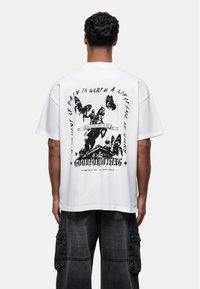 Biała, oversize'owa koszulka z grafiką, z czarnym nadrukiem przedstawiającym motyle i postacie, okrągły dekolt i krótkie rękawy. Design na plecach zawiera tekst.