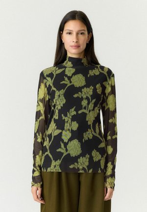 Kvinde iført en tynd sort rullekravetrøje med grøn floral broderi og olivengrønne bukser, stående mod en ensfarvet lys baggrund.