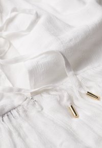 Tessuto bianco con un motivo testurizzato, dotato di cordini con punte metalliche dorate e una leggera arricciatura elastica in fondo.
