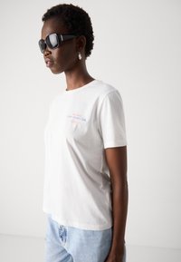 T-shirt bianca in cotone con maniche corte e scollatura rotonda, caratterizzata da testo blu e rosso sulla parte anteriore. Indossata con jeans azzurri chiari.