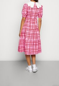 Femme portant une robe midi à carreaux rose et rouge avec des manches bouffantes, un grand col blanc et des baskets montantes blanches, se tenant contre un mur uni.