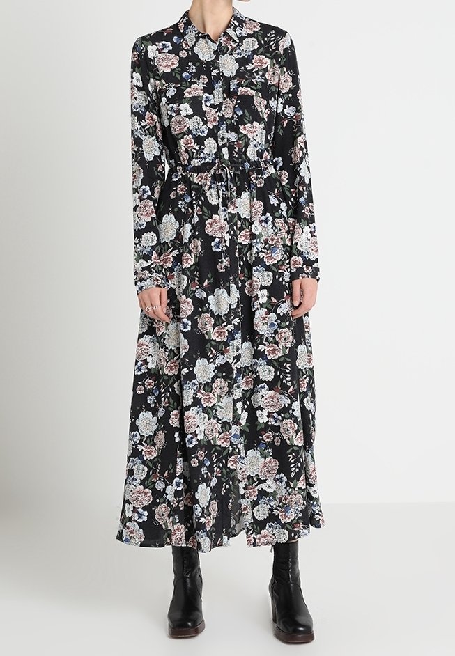 Femme portant une robe maxi noire à fleurs à manches longues, avec une taille à cordon et des bottines noires.