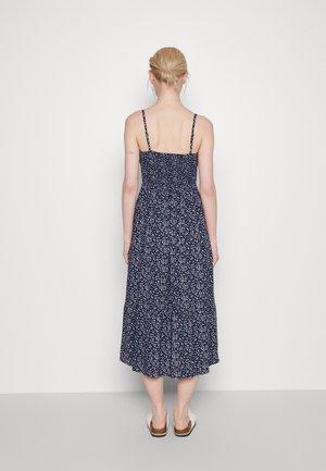 Kvinde med blondt hår sat i en lav knold, iført en marineblå blomsterprintet midikjole med tynde stropper, stående barfodet mod en ensfarvet hvid baggrund.