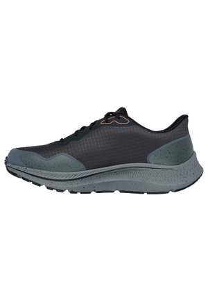 Zapatilla deportiva negra con parte superior texturizada, suela moteada en gris y detalles en naranja contrastantes. Presenta un diseño de cordones y punta redondeada.