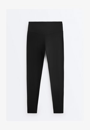 Zwarte legging met hoge taille, een naadloze tailleband, taps toelopende pijpen en glad, rekbaar materiaal tegen een effen witte achtergrond.