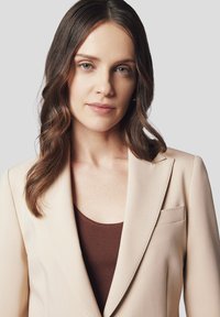 Beżowa marynarka z gładkim kołnierzem i detalami kieszeni, noszona na brązowej, prążkowanej bluzce. Gładki materiał o dopasowanym kroju, z delikatnym przeszyciem.