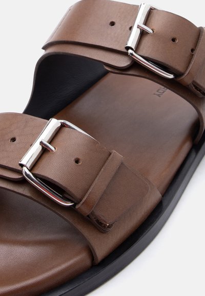 Καφέ δερμάτινο σανδάλι με δύο ρυθμιζόμενες ασημένιες αγκράφες και μαύρη σόλα, που δείχνει λεπτομερή υφή και ραφές στα λουριά.