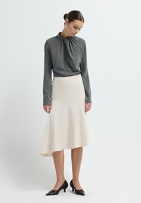 Grijze langsluisse top met geplooide halslijn, gecombineerd met een crèmekleurige uitlopende rok met een asymmetrische zoom en zwarte puntige hakken.