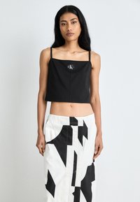 Sort cropped topp med tynne stropper og et merke med "CK." Kombinert med et høyt liv hvitt skjørt som viser svarte geometriske mønstre.