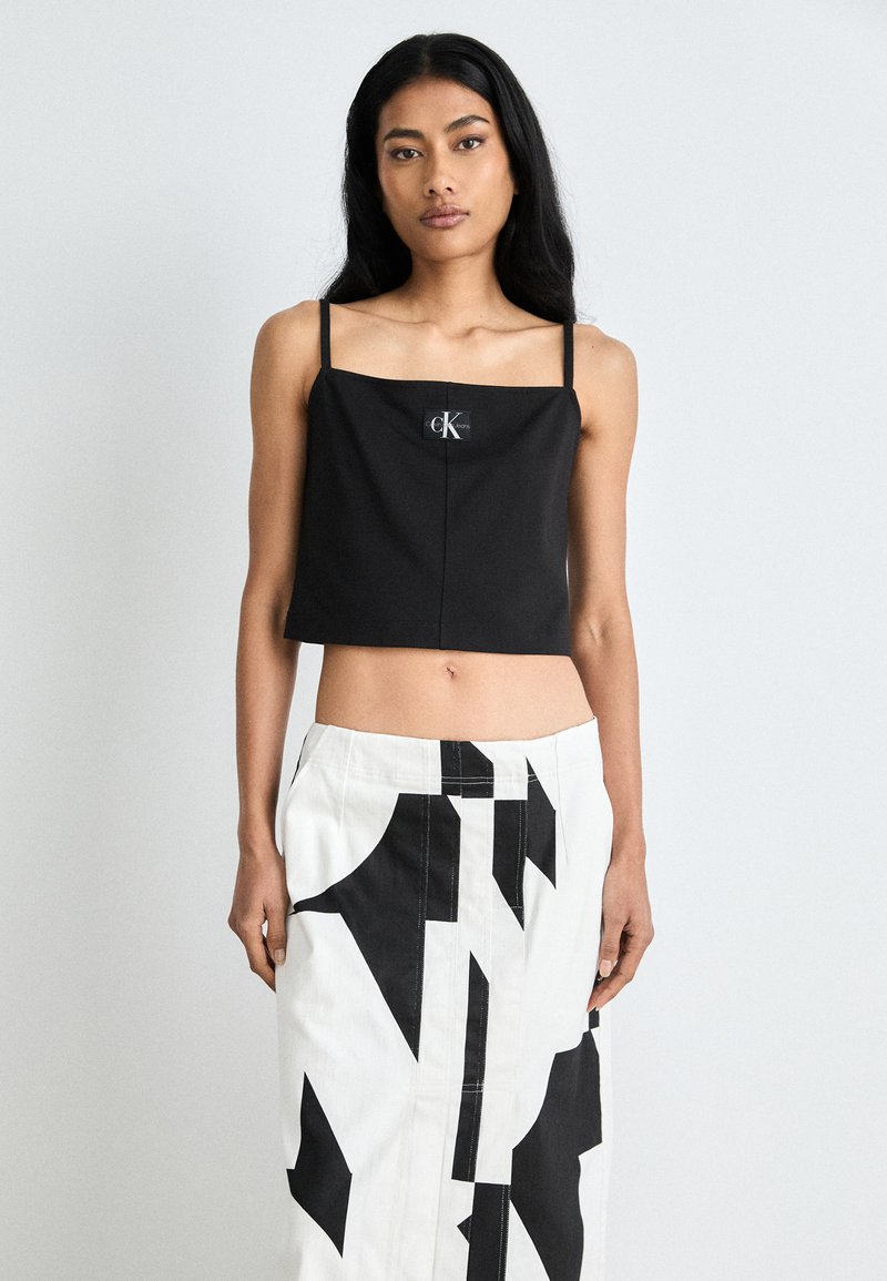 Sort cropped topp med tynne stropper og et merke med "CK." Kombinert med et høyt liv hvitt skjørt som viser svarte geometriske mønstre.