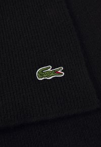 Čierna rib omotaná tkanina s malým zeleným vyšitým logom krokodíla s červenými detailmi a bielym obrysom.