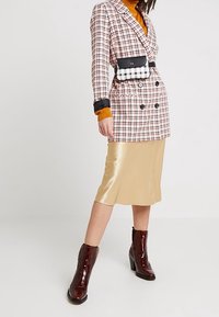 Blazer a quadri rosso e bianco con design doppiopetto, abbinato a una gonna in satin oro e stivaletti lucidi marroni alla caviglia.