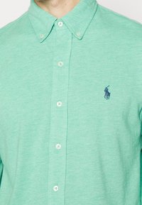 Man wearing green button-up shirt with collar and dark embroidered polo player logo on chest.