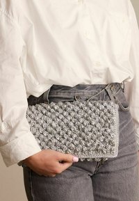 Pochette argentata, con texture e motivo a pallini, di forma rettangolare, tenuta in una mano contro una camicia bianca e jeans grigi.