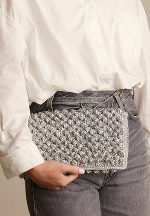 Sac à main texturé argenté avec un motif en boules, de forme rectangulaire, tenu d'une main contre une chemise blanche et un jean gris.