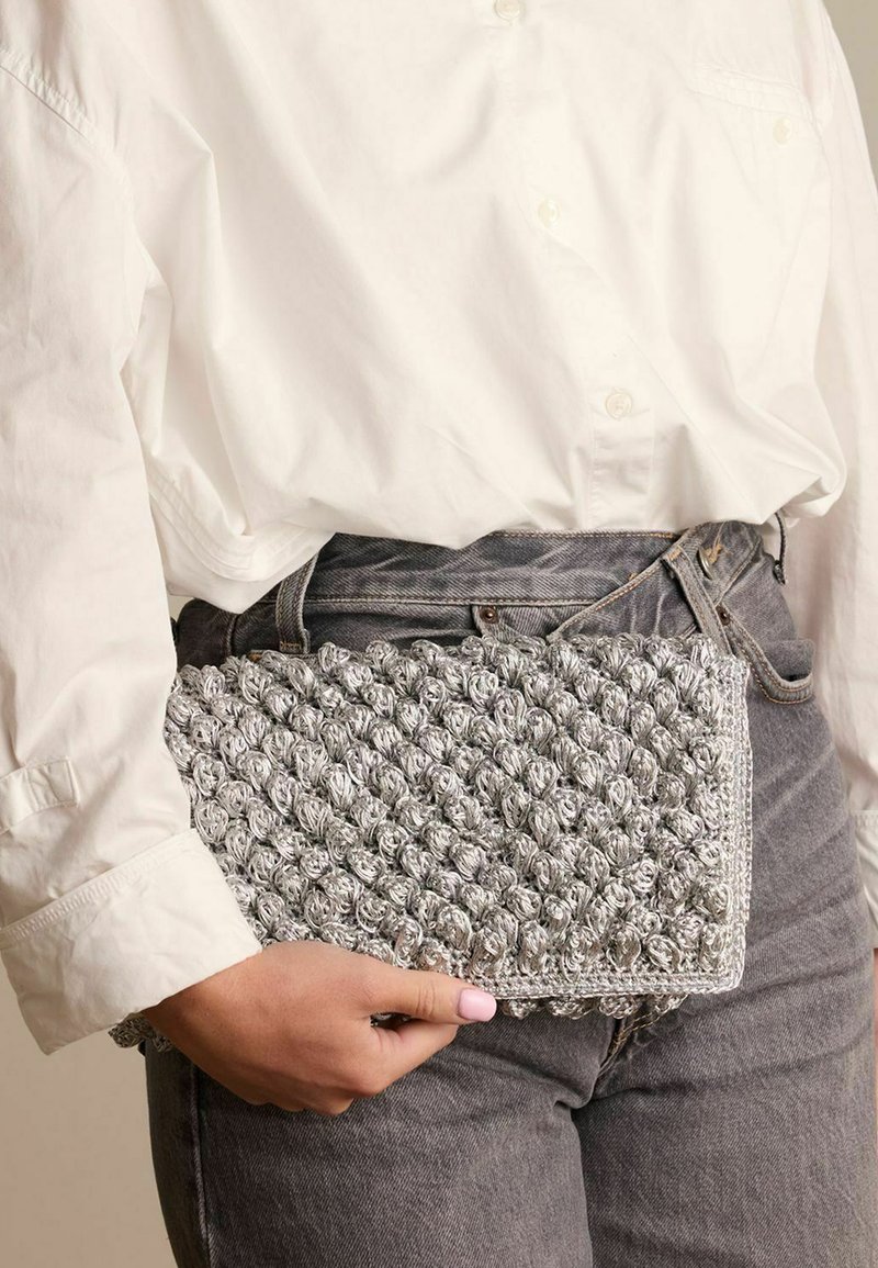 Pochette argentata, con texture e motivo a pallini, di forma rettangolare, tenuta in una mano contro una camicia bianca e jeans grigi.