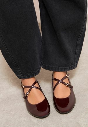 Voeten met donker bordeauxrode Mary Jane-lakleren schoenen met dubbele bandjes en gouden gespen, gecombineerd met zwarte wijde broekspijpen.