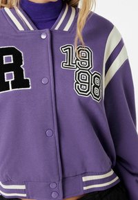 Lilla cropped varsity jakke med hvidstribede kraver, prydet med broderede bogstaver i sort og hvid "R" samt "1998" patches, og trykknapper.