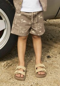 Un short marron clair avec un motif de paysage désertique, associé à des sandales beiges à deux straps réglables, posées sur un sol sablonneux.