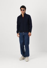 Laikina jūrų laivyno zip-up striukė su aukštu apykakle, banguotais rankogaliais ir dviem priekinių kišenių, dėvima ant bordo marškinėlių ir derinama su mėlynomis džinsomis bei smėlio spalvos batais.