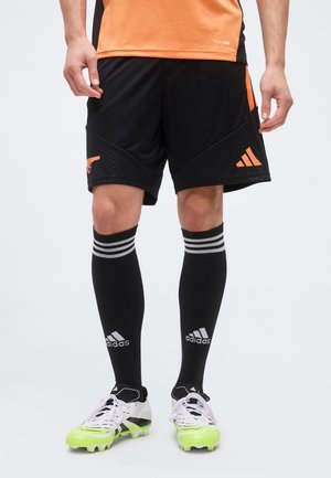 Mand iført sorte og orange fodboldshorts, sorte Adidas knæstrømper og hvide fodboldstøvler med neongule såler på en ensfarvet baggrund.