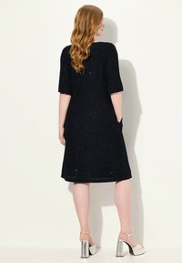 Robe noire mi-longue avec une texture pailletée, manches courtes et poches latérales ; portée avec des talons compensés en argent brillants.