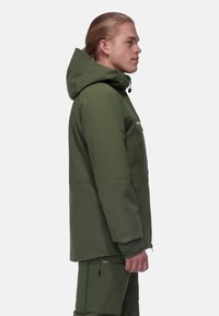Giacca impermeabile verde con cappuccio, tasche con zip e finitura opaca. Presenta una vestibilità rilassata e maniche articolate.