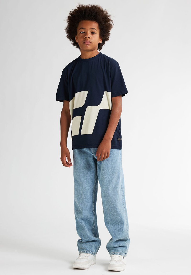 Niño con peinado afro vestido con camiseta azul marino con estampado geométrico blanco, vaqueros azul claro y zapatillas blancas, sobre un fondo blanco.