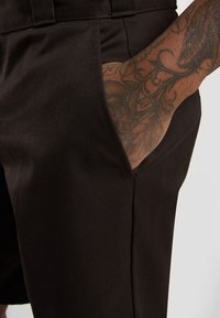Black tailored shorts with a smooth texture, featuring a side pocket and a close-up of a detailed tattooed arm.