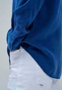 Blaues Langarm-Hemd mit Knopfleiste, glatter Textur und aufgerollten Ärmeln, kombiniert mit weißen Denim-Hosen, die über eine Seitentasche verfügen.