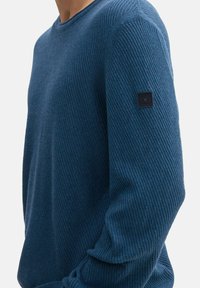 Maglione blu con texture e motivo a coste, colletto rotondo e una patch nera con logo sulla manica sinistra. Realizzato in materiale morbido.