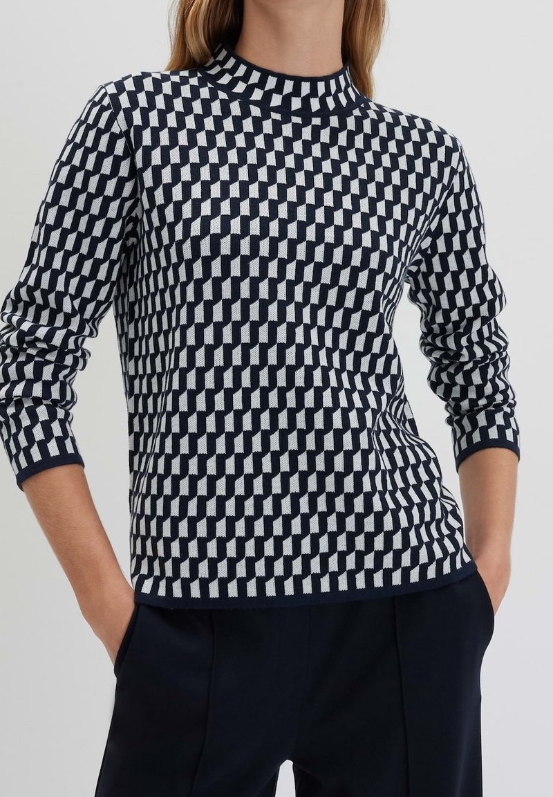 Personne portant un pull à manches longues avec un motif à carreaux géométrique noir et blanc et un pantalon foncé, les mains dans les poches.