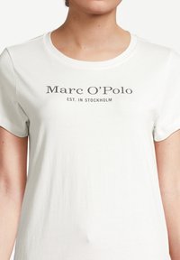 Vit bomulls t-shirt med rund hals och korta ärmar. Har "Marc O'Polo EST. I STOCKHOLM" tryckt i grått på framsidan.