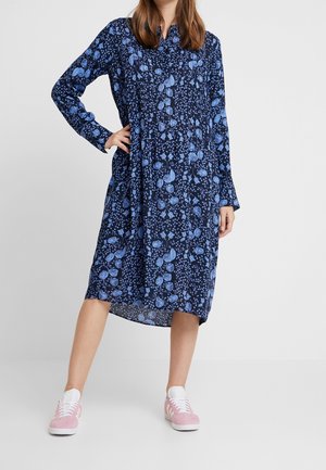 Kvinde iført en blå blomsterprint midi-kjole med lange ærmer og lyserøde og hvide sneakers, stående med den ene hånd på hoften mod en grå baggrund.