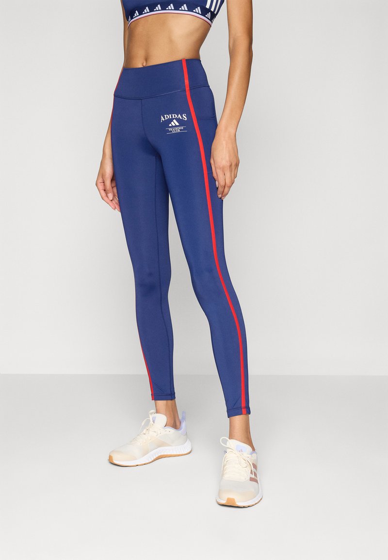 Marineblauwe leggings met rode zijstrepen, hoge taille, gladde textuur en witte "ADIDAS" logo detail. Draagt met witte en grijze sportschoenen.